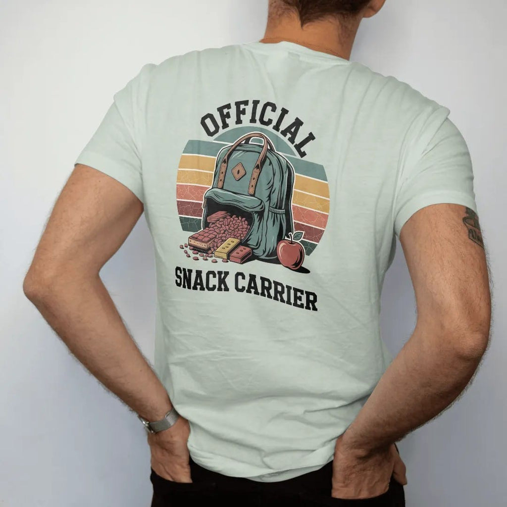 Man wearing a Bay tee, seen from the back: a bulging daypack spilling trail mix, granola bars and an apple, merit-badge layout, faded 70s park-souvenir style, lettered "OFFICIAL / SNACK CARRIER" in chunky rounded 70s serif letters with a drop…