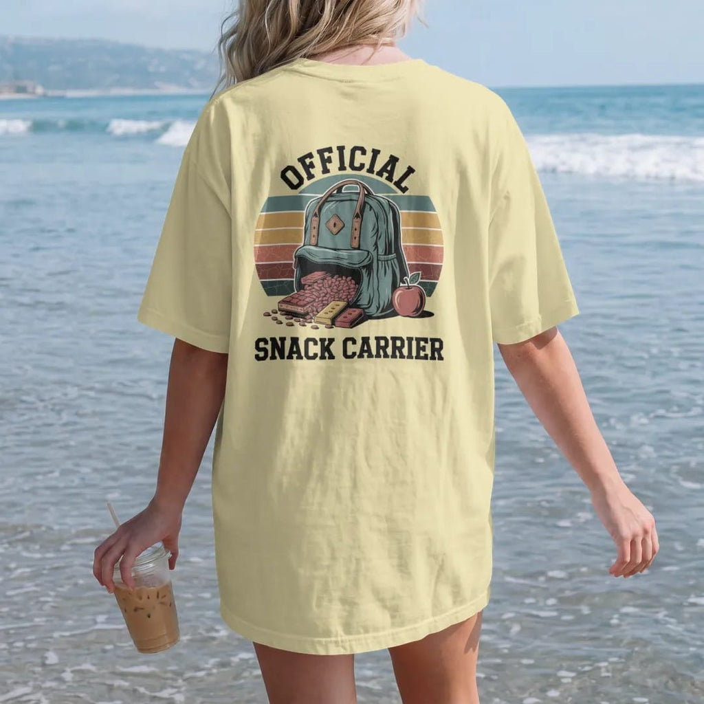 Woman on the beach in a Butter tee, back view: a bulging daypack spilling trail mix, granola bars and an apple, merit-badge layout, faded 70s park-souvenir style, lettered "OFFICIAL / SNACK CARRIER" in chunky rounded 70s serif letters with a drop…