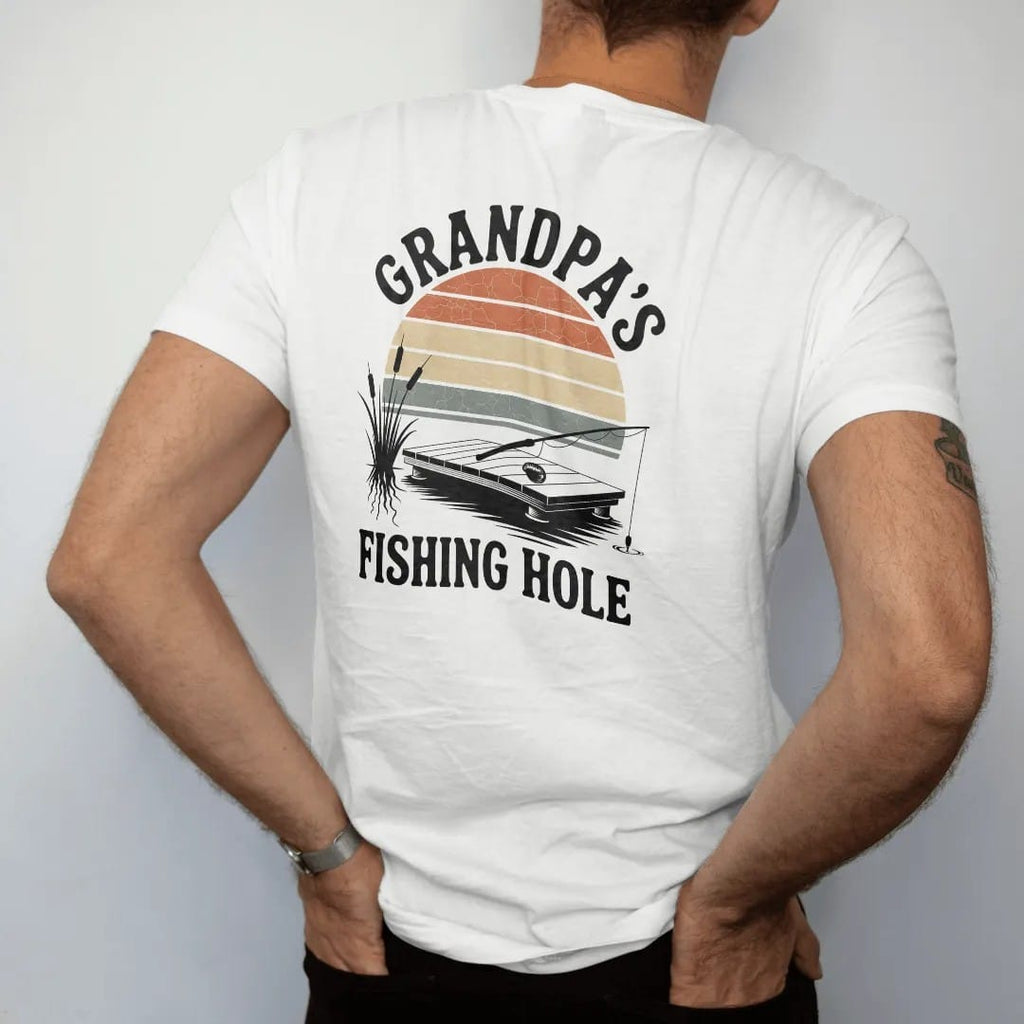 Man wearing a White tee, seen from the back: a small wooden dock on a quiet pond with a bobber, cattails and a resting fishing rod, faded 70s park-souvenir style, lettered "GRANDPA'S / FISHING HOLE" in chunky rounded 70s letters with a drop shadow,…