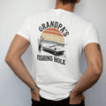 Man wearing a White tee, seen from the back: a small wooden dock on a quiet pond with a bobber, cattails and a resting fishing rod, faded 70s park-souvenir style, lettered "GRANDPA'S / FISHING HOLE" in chunky rounded 70s letters with a drop shadow,…