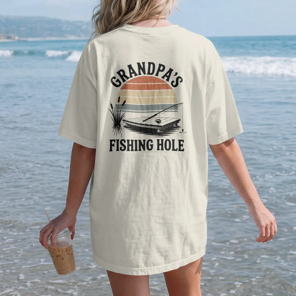 Woman on the beach in a Ivory tee, back view: a small wooden dock on a quiet pond with a bobber, cattails and a resting fishing rod, faded 70s park-souvenir style, lettered "GRANDPA'S / FISHING HOLE" in chunky rounded 70s letters with a drop…