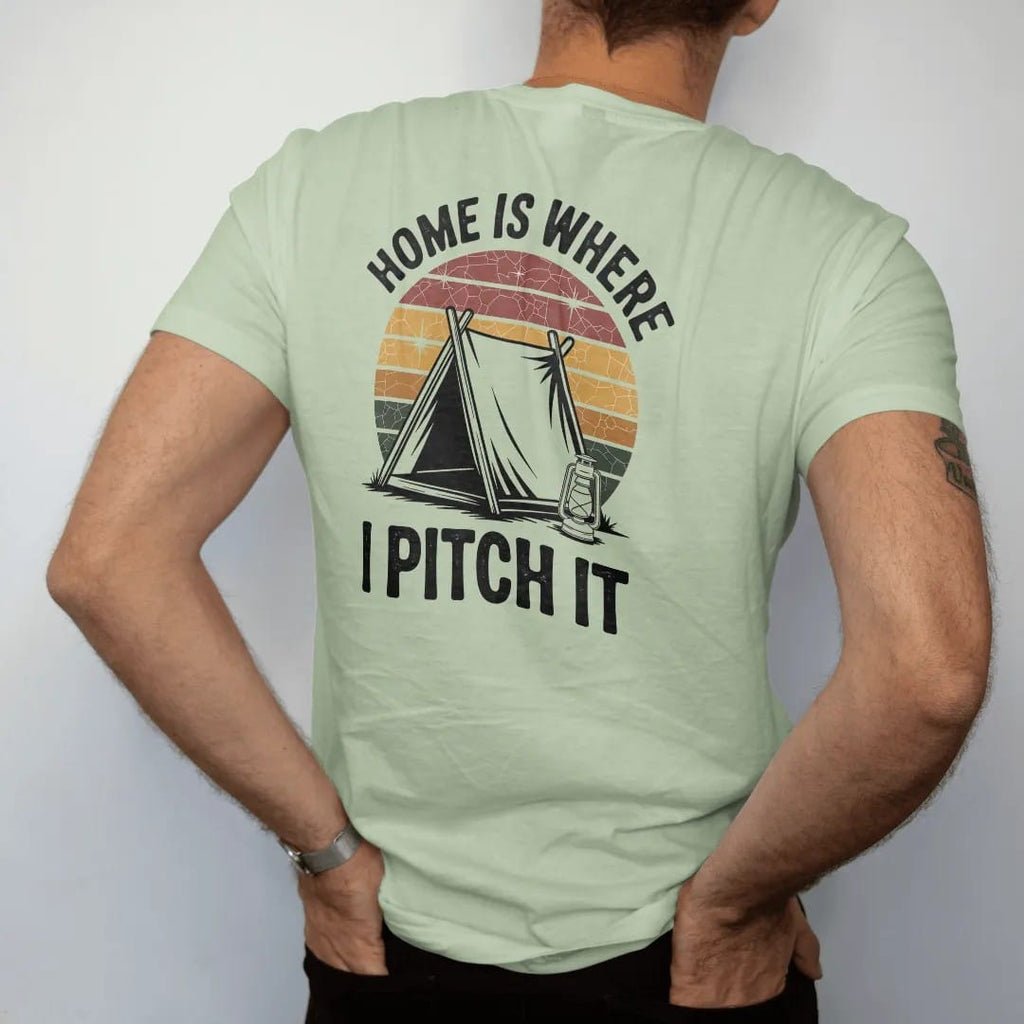 Man wearing a Light Green tee, seen from the back: an A-frame tent pitched under a starry sky beside a little lantern, stacked text above, faded 70s park-souvenir style, lettered "HOME IS WHERE / I PITCH IT" in chunky rounded 70s letters with a…
