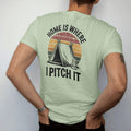 Man wearing a Light Green tee, seen from the back: an A-frame tent pitched under a starry sky beside a little lantern, stacked text above, faded 70s park-souvenir style, lettered "HOME IS WHERE / I PITCH IT" in chunky rounded 70s letters with a…