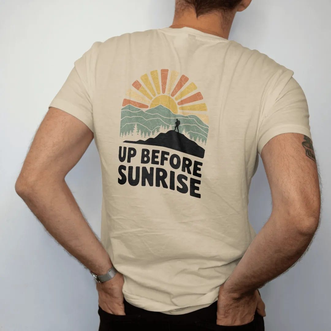 Man wearing a Sandstone tee, seen from the back: a sun peeking over layered mountain ridges with long rays and a tiny hiker on a ridge, faded 70s park-souvenir style, lettered "UP BEFORE SUNRISE" in chunky rounded 70s letters with a drop shadow,…