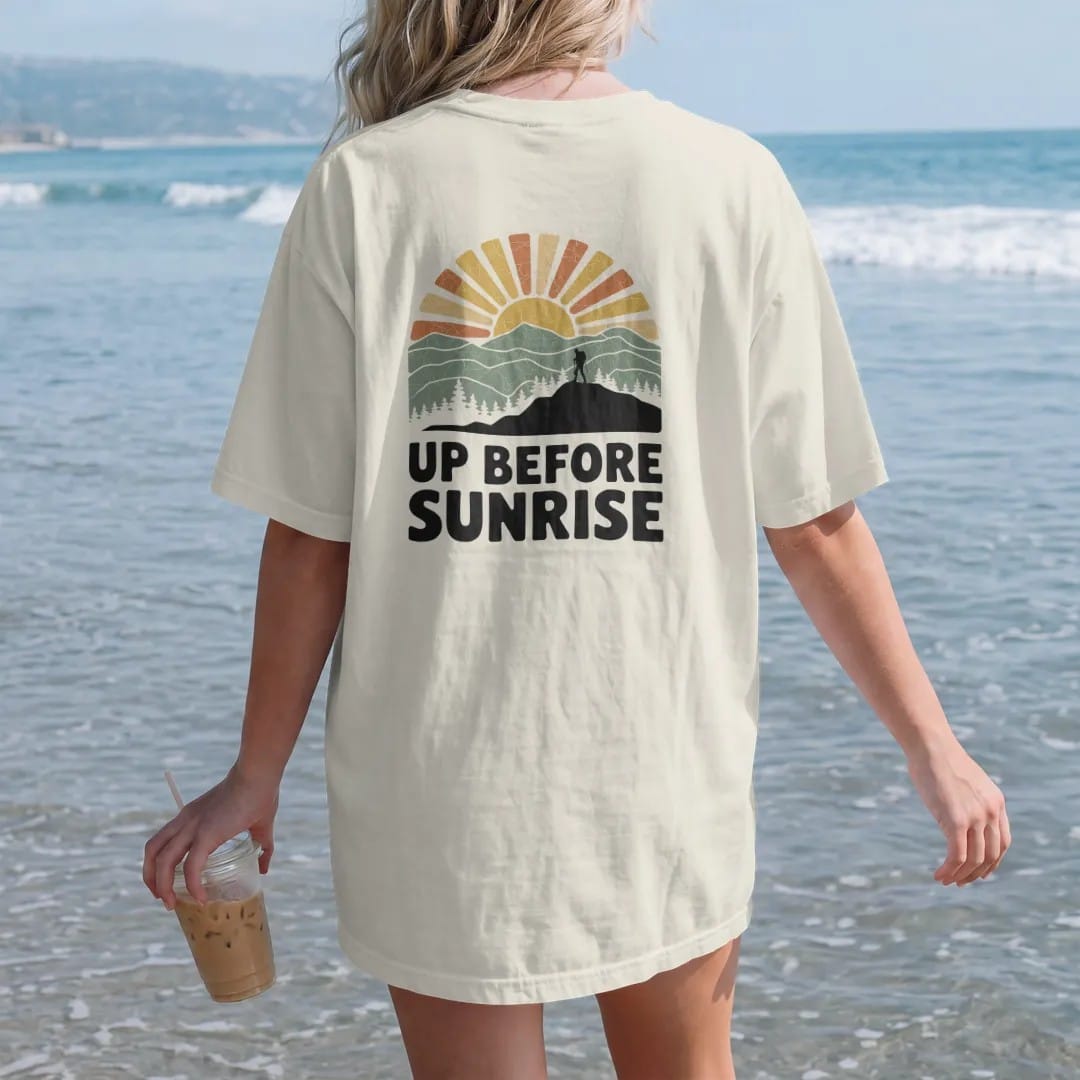 Woman on the beach in a Ivory tee, back view: a sun peeking over layered mountain ridges with long rays and a tiny hiker on a ridge, faded 70s park-souvenir style, lettered "UP BEFORE SUNRISE" in chunky rounded 70s letters with a drop shadow,…