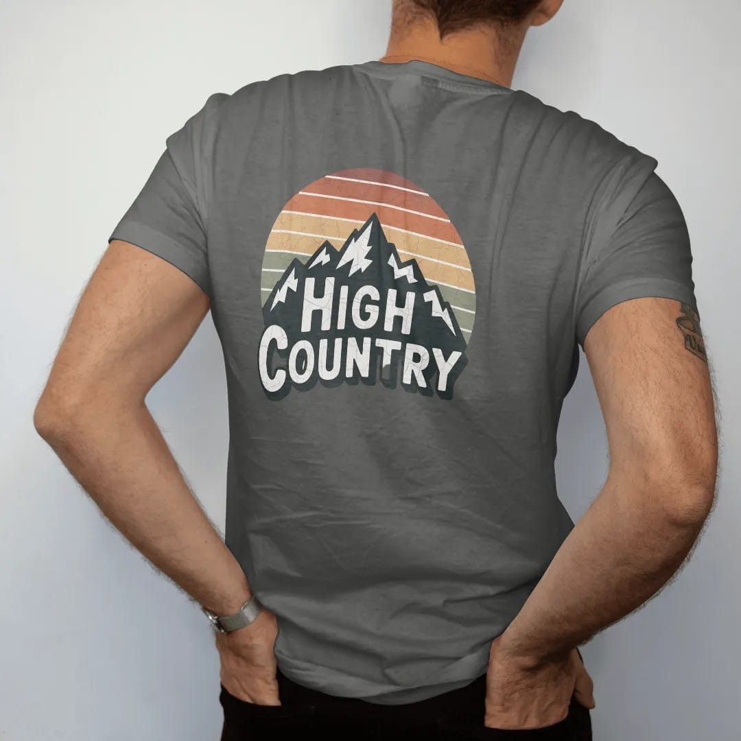 Man wearing a Pepper tee, seen from the back: a sweeping mountain range wordmark with snowcaps, the letters sitting on the ridgeline, faded 70s park-souvenir style, lettered "HIGH COUNTRY" in chunky rounded 70s letters with a drop shadow, printed…