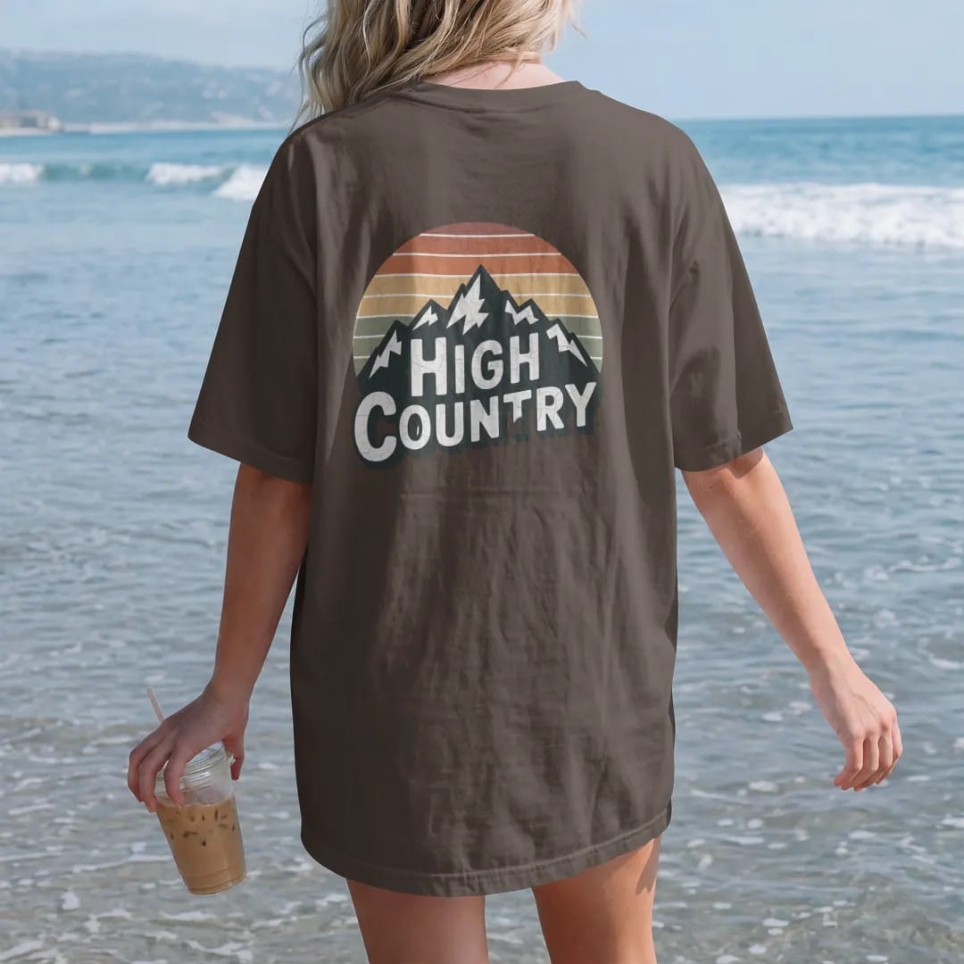 Woman on the beach in a Espresso tee, back view: a sweeping mountain range wordmark with snowcaps, the letters sitting on the ridgeline, faded 70s park-souvenir style, lettered "HIGH COUNTRY" in chunky rounded 70s letters with a drop shadow,…