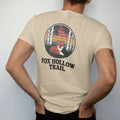 Man wearing a Sandstone tee, seen from the back: a red fox curled up in tall grass under birch trees inside a round patch frame, faded 70s park-souvenir style, lettered "FOX HOLLOW TRAIL" in chunky rounded 70s serif letters with a drop shadow,…