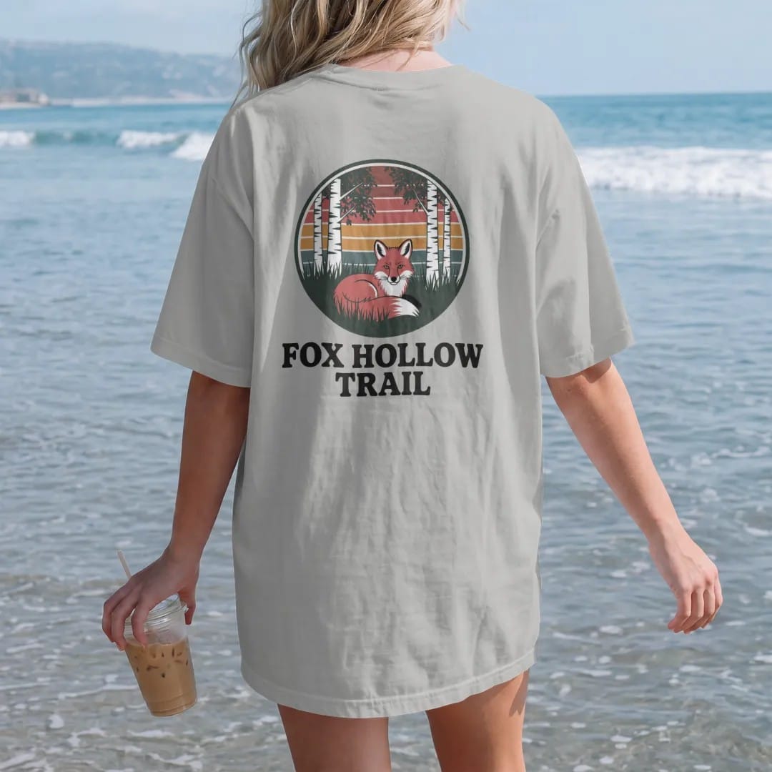 Woman on the beach in a Granite tee, back view: a red fox curled up in tall grass under birch trees inside a round patch frame, faded 70s park-souvenir style, lettered "FOX HOLLOW TRAIL" in chunky rounded 70s serif letters with a drop shadow,…