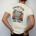 Man wearing a Ivory tee, seen from the back: a cluster of illustrated wild mushrooms and ferns in a woven basket, field-guide layout, faded 70s park-souvenir style, lettered "FORAGE CLUB / ASK FIRST" in chunky rounded 70s serif letters with a drop…