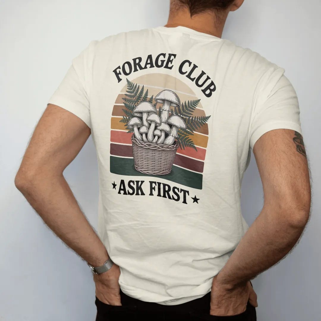 Man wearing a Ivory tee, seen from the back: a cluster of illustrated wild mushrooms and ferns in a woven basket, field-guide layout, faded 70s park-souvenir style, lettered "FORAGE CLUB / ASK FIRST" in chunky rounded 70s serif letters with a drop…
