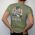 Man wearing a Moss tee, seen from the back: a row of tall tree trunks each painted with a trail blaze rectangle, path winding between them, faded 70s park-souvenir style, lettered "FOLLOW THE BLAZES" in chunky rounded 70s letters with a drop…