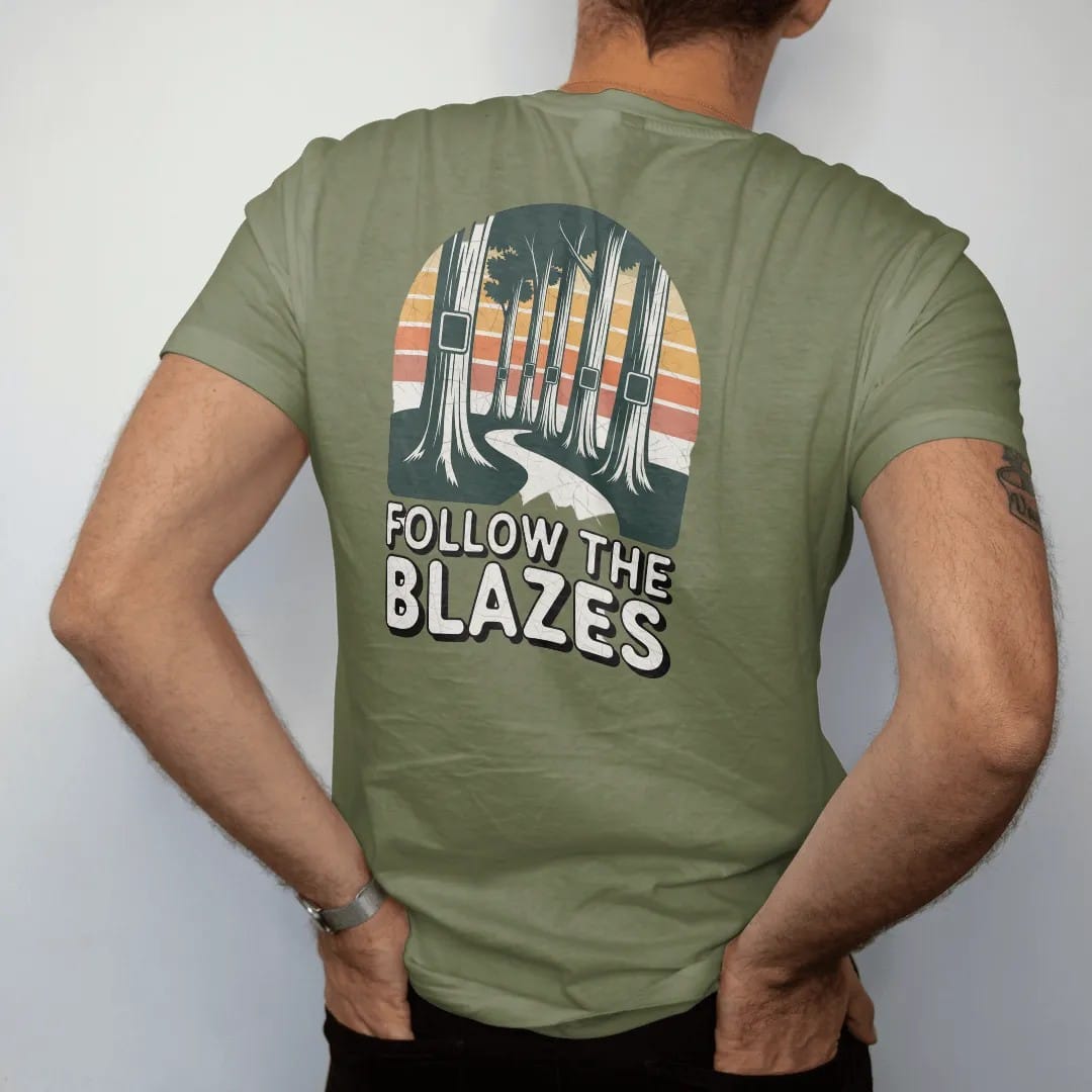 Man wearing a Moss tee, seen from the back: a row of tall tree trunks each painted with a trail blaze rectangle, path winding between them, faded 70s park-souvenir style, lettered "FOLLOW THE BLAZES" in chunky rounded 70s letters with a drop…