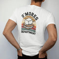 Man wearing a White tee, seen from the back: a toasted marshmallow on a stick dripping between graham crackers, official-department seal layout, faded 70s park-souvenir style, lettered "S'MORES / DEPARTMENT" in chunky rounded 70s serif letters with…