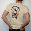 Man wearing a Sandstone tee, seen from the back: an old kerosene camp lantern glowing on a stump with moths and stars, warm light rays, faded 70s park-souvenir style, lettered "LANTERN LIGHT / CAMP" in chunky rounded 70s serif letters with a drop…