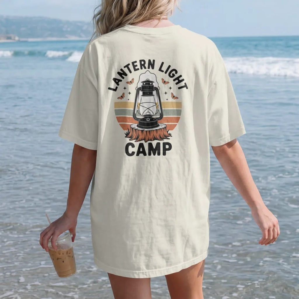 Woman on the beach in a Ivory tee, back view: an old kerosene camp lantern glowing on a stump with moths and stars, warm light rays, faded 70s park-souvenir style, lettered "LANTERN LIGHT / CAMP" in chunky rounded 70s serif letters with a drop…