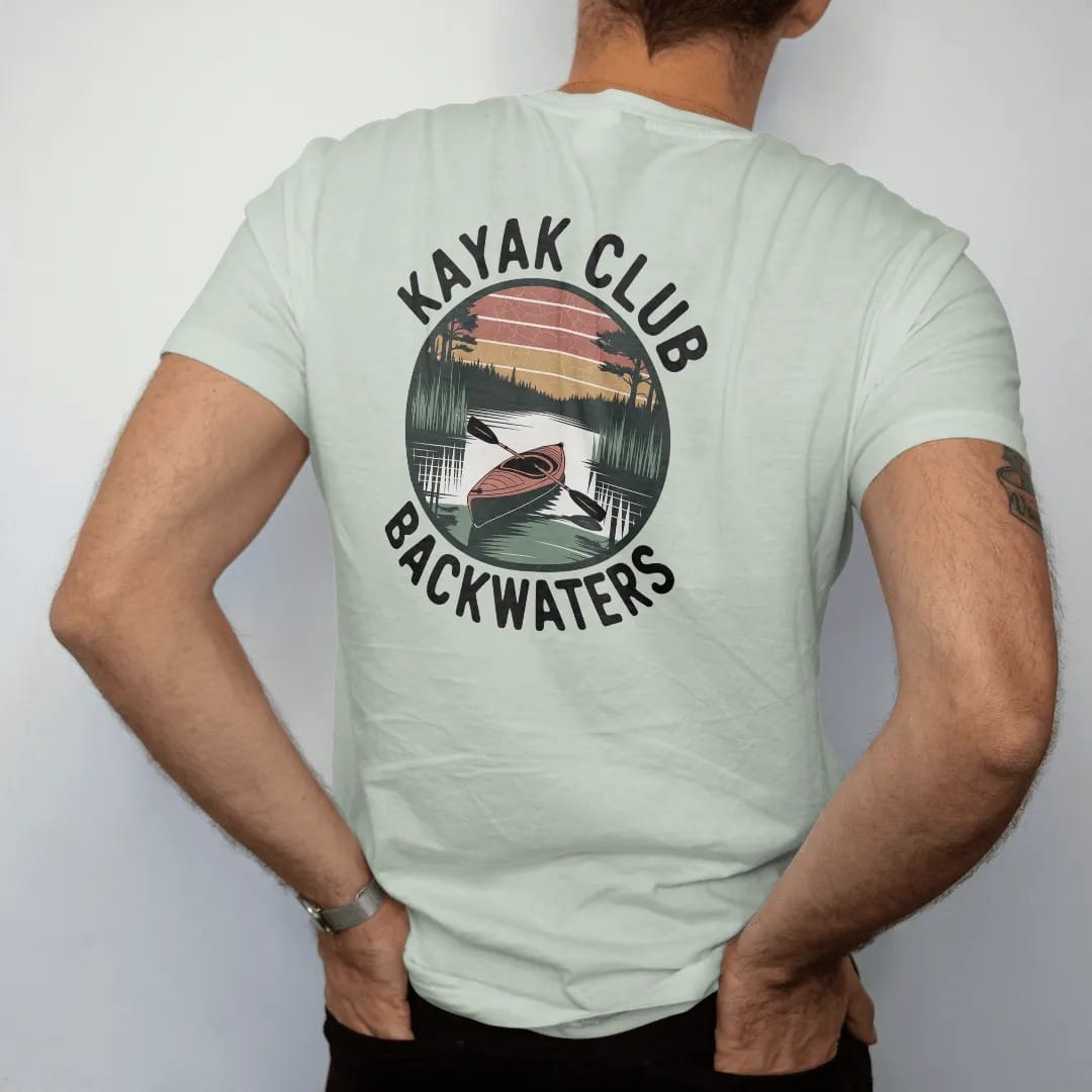 Man wearing a Bay tee, seen from the back: a kayak gliding through a cypress swamp with herons and calm reflections, round badge frame, faded 70s park-souvenir style, lettered "KAYAK CLUB / BACKWATERS" in chunky rounded 70s letters with a drop…