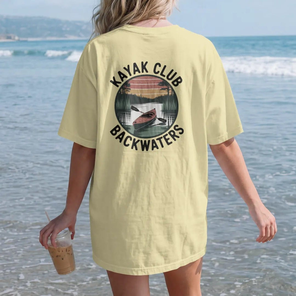 Woman on the beach in a Butter tee, back view: a kayak gliding through a cypress swamp with herons and calm reflections, round badge frame, faded 70s park-souvenir style, lettered "KAYAK CLUB / BACKWATERS" in chunky rounded 70s letters with a drop…