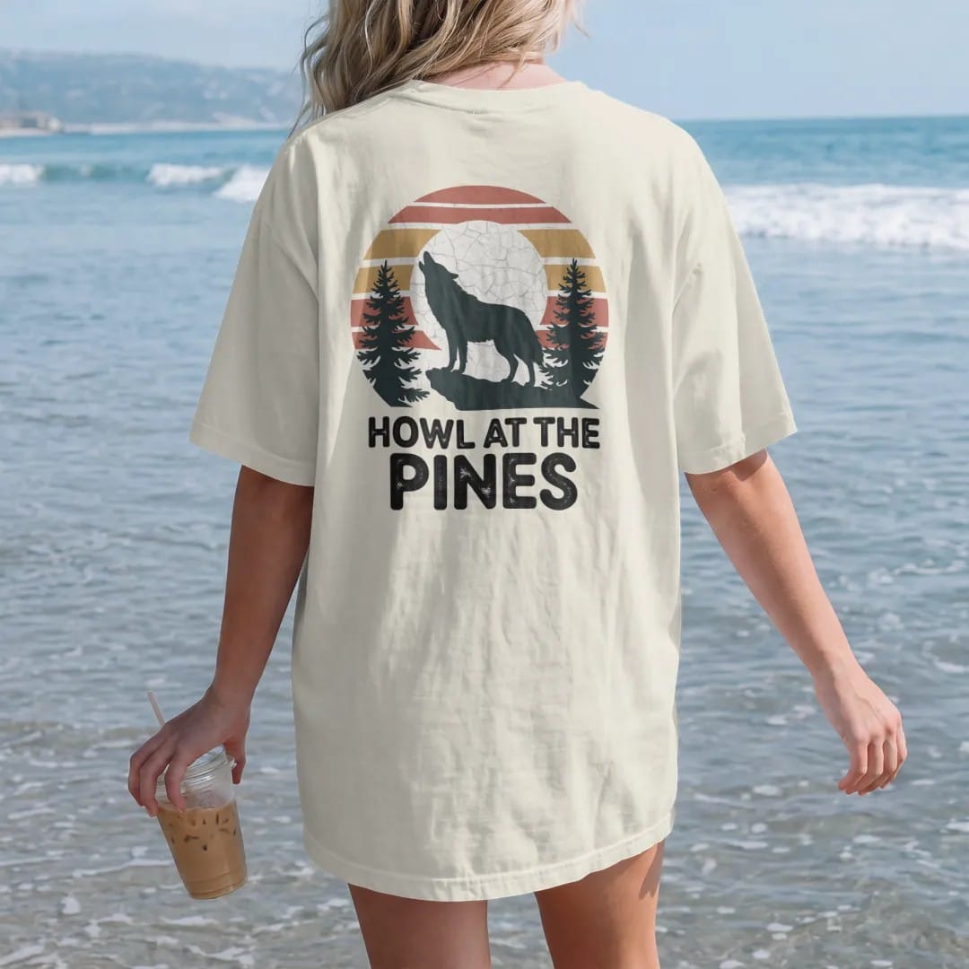Woman on the beach in a Ivory tee, back view: a wolf howling on a rocky ledge with a full moon behind it and pines in silhouette, faded 70s park-souvenir style, lettered "HOWL AT THE PINES" in chunky rounded 70s letters with a drop shadow, printed…