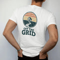 Man wearing a White tee, seen from the back: a circular topo-line badge with a tiny cabin at the center and a dotted trail leading out, faded 70s park-souvenir style, lettered "OFF THE GRID" in chunky rounded 70s serif letters with a drop shadow,…