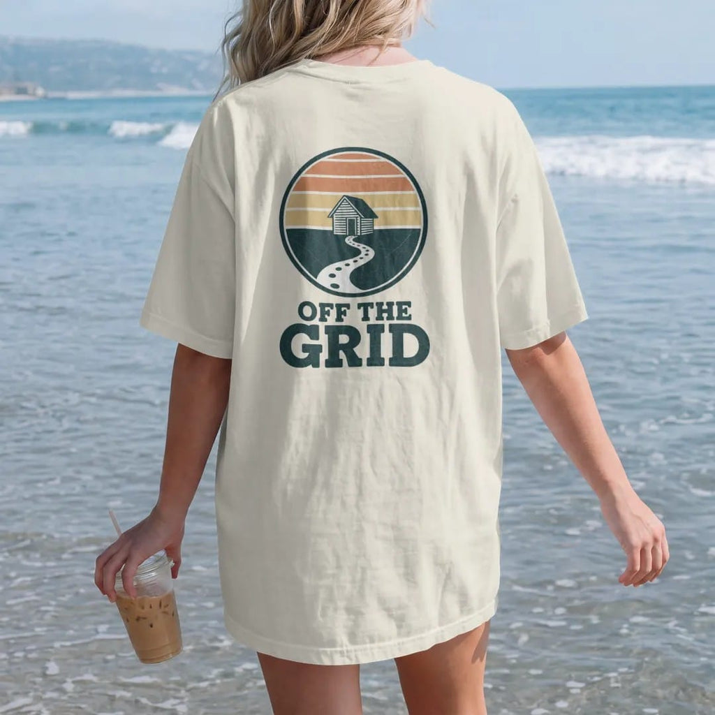 Woman on the beach in a Ivory tee, back view: a circular topo-line badge with a tiny cabin at the center and a dotted trail leading out, faded 70s park-souvenir style, lettered "OFF THE GRID" in chunky rounded 70s serif letters with a drop shadow,…