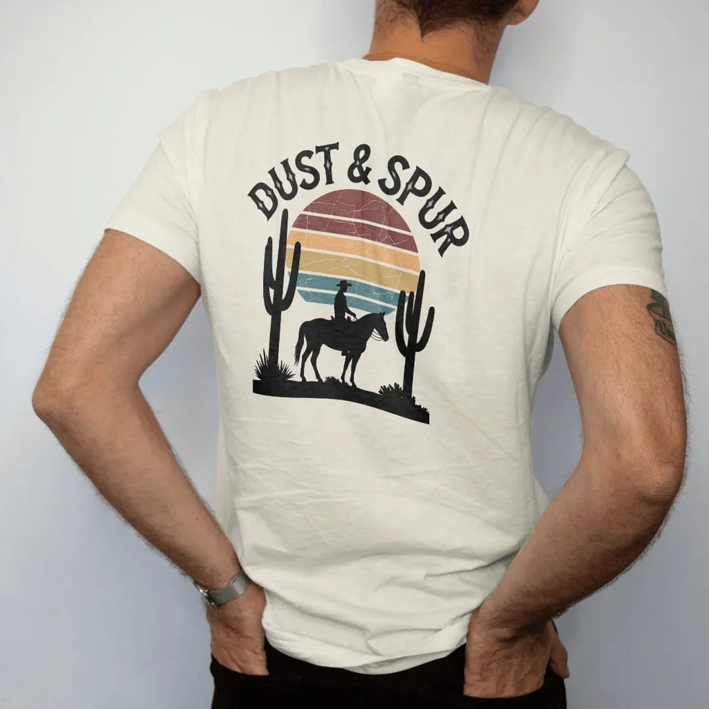Man wearing a Ivory tee, seen from the back: a lone cowboy on horseback silhouetted against a big retro-striped desert sun with saguaros on either side, "DUST & SPUR" in a faded arched Tuscan rodeo-poster header above, in a faded 1970s souvenir…