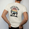 Man wearing a Ivory tee, seen from the back: a lone cowboy on horseback silhouetted against a big retro-striped desert sun with saguaros on either side, "DUST & SPUR" in a faded arched Tuscan rodeo-poster header above, in a faded 1970s souvenir…