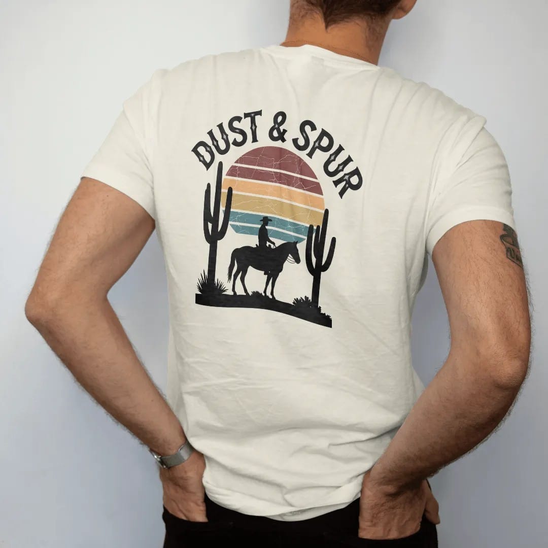 Man wearing a Ivory tee, seen from the back: a lone cowboy on horseback silhouetted against a big retro-striped desert sun with saguaros on either side, "DUST & SPUR" in a faded arched Tuscan rodeo-poster header above, in a faded 1970s souvenir…