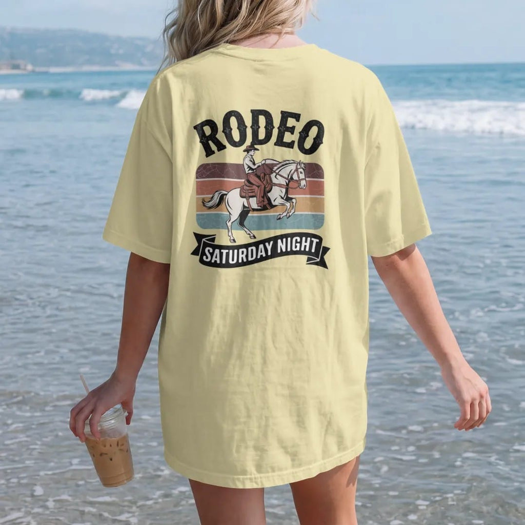 Woman on the beach in a Butter tee, back view: vintage rodeo poster layout with a bucking bronco and rider, "RODEO" in huge chunky 70s slab letters above and "SATURDAY NIGHT" on a banner below, in a faded 1970s souvenir style with a worn…