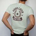 Man wearing a Bay tee, seen from the back: a worn horseshoe with nail holes framing a small eight-point star, "GOOD LUCK" arched above in chunky 70s block letters and "RIDE HARD" on a ribbon below, in a faded 1970s souvenir style with a worn…