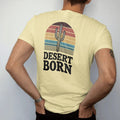 Man wearing a Butter tee, seen from the back: a tall saguaro cactus with two arms in front of a sliced half sun with retro rainbow bands and distant mesas, "DESERT BORN" in chunky 70s block letters below, in a faded 1970s souvenir style with a worn…