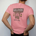 Man wearing a Watermelon tee, seen from the back: an old wooden saloon sign with batwing doors beneath it and two hanging lanterns, "SALOON" in faded Tuscan rodeo-poster letters and "OPEN LATE" painted small below, in a faded 1970s souvenir style…