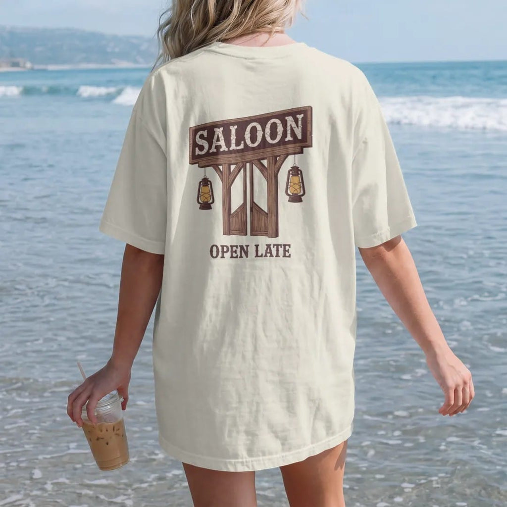 Woman on the beach in a Ivory tee, back view: an old wooden saloon sign with batwing doors beneath it and two hanging lanterns, "SALOON" in faded Tuscan rodeo-poster letters and "OPEN LATE" painted small below, in a faded 1970s souvenir style with…