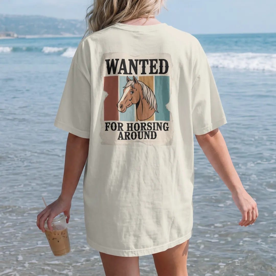 Woman on the beach in a Ivory tee, back view: an old torn wanted poster with a cartoon horse portrait in the center, "WANTED" in chunky 70s slab letters at top and "FOR HORSING AROUND" in small caps below, in a faded 1970s souvenir style with a…