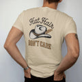 Man wearing a Sandstone tee, seen from the back: a worn cowboy hat with a stitched band and a single feather, "HAT HAIR" in a looping 70s rope script above and "DON'T CARE" in chunky 70s block letters below, in a faded 1970s souvenir style with a…