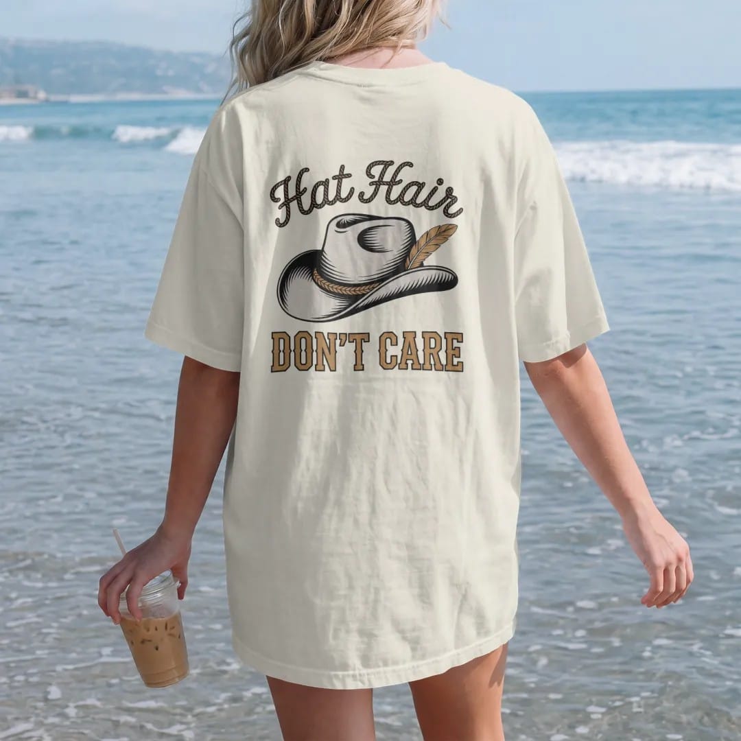 Woman on the beach in a Ivory tee, back view: a worn cowboy hat with a stitched band and a single feather, "HAT HAIR" in a looping 70s rope script above and "DON'T CARE" in chunky 70s block letters below, in a faded 1970s souvenir style with a worn…