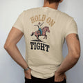 Man wearing a Sandstone tee, seen from the back: a bronc rider mid-buck with one hand high and dust kicking up, "HOLD ON" in chunky 70s slab letters above and "TIGHT" below, in a faded 1970s souvenir style with a worn cracked-ink texture, printed…
