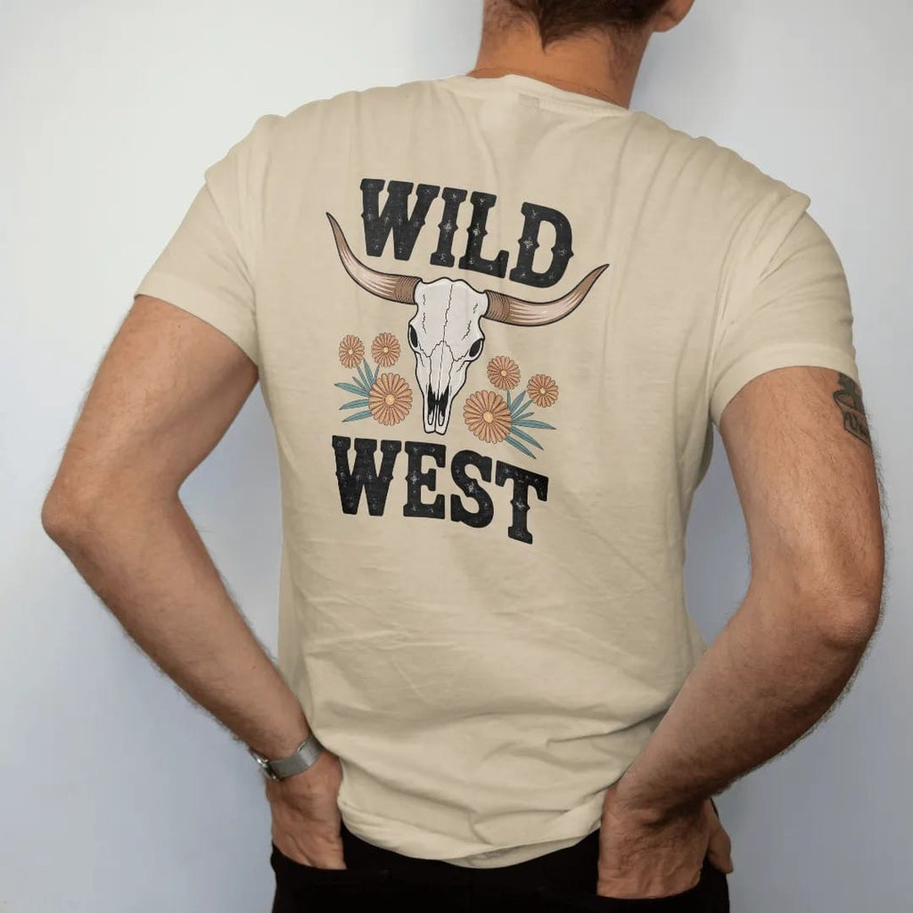 Man wearing a Sandstone tee, seen from the back: a sun-bleached longhorn steer skull with wide curved horns and a few desert wildflowers below, "WILD WEST" in chunky 70s block letters beneath, in a faded 1970s souvenir style with a worn cracked-ink…
