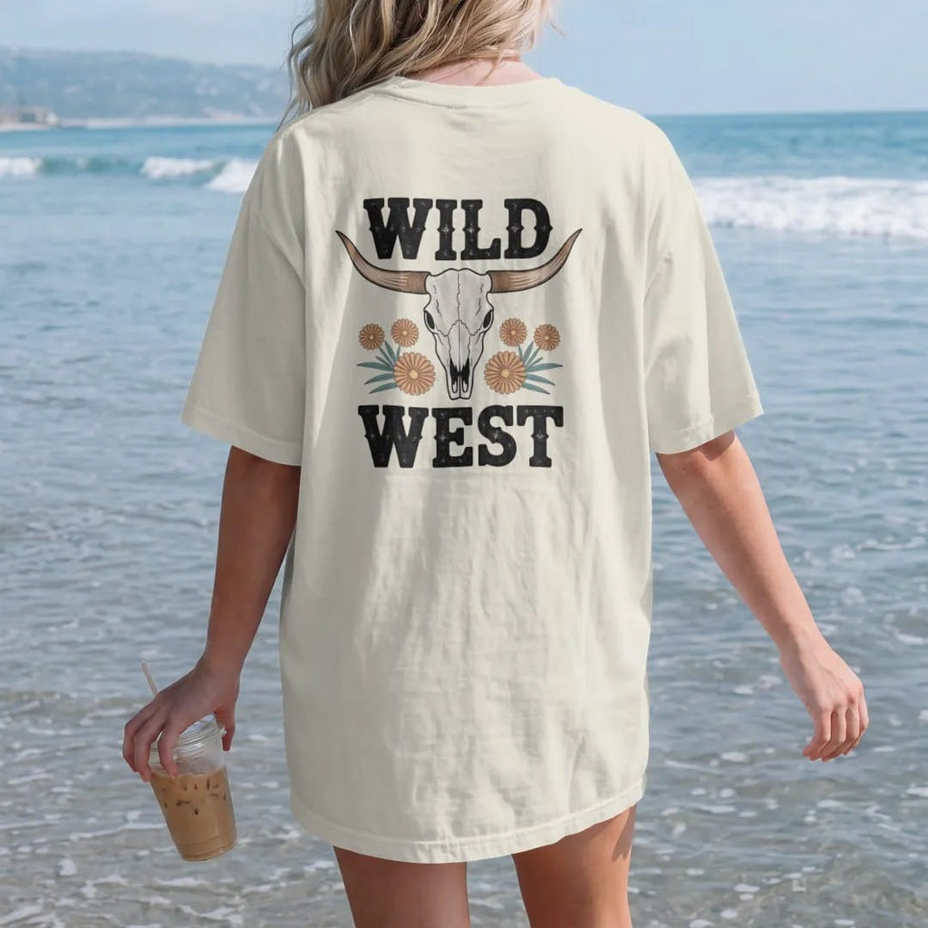Woman on the beach in a Ivory tee, back view: a sun-bleached longhorn steer skull with wide curved horns and a few desert wildflowers below, "WILD WEST" in chunky 70s block letters beneath, in a faded 1970s souvenir style with a worn cracked-ink…