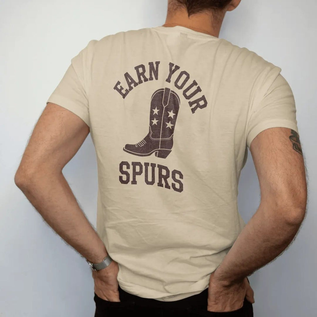 Man wearing a Sandstone tee, seen from the back: a single cowboy boot spur with a star rowel, "EARN YOUR SPURS" in an arched header of chunky 70s slab letters above, in a faded 1970s souvenir style with a worn cracked-ink texture, printed in saddle…