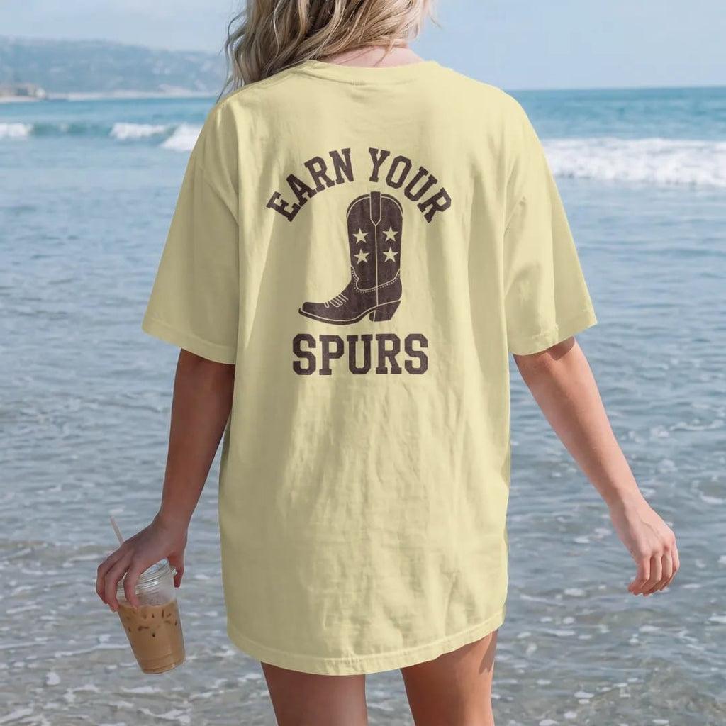 Woman on the beach in a Butter tee, back view: a single cowboy boot spur with a star rowel, "EARN YOUR SPURS" in an arched header of chunky 70s slab letters above, in a faded 1970s souvenir style with a worn cracked-ink texture, printed in saddle…