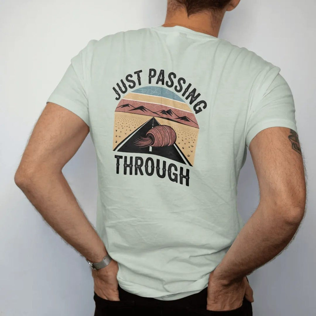Man wearing a Bay tee, seen from the back: a tumbleweed rolling across a flat desert road toward far mountains, "JUST PASSING THROUGH" in small retro 70s caps beneath, in a faded 1970s souvenir style with a worn cracked-ink texture, printed in…