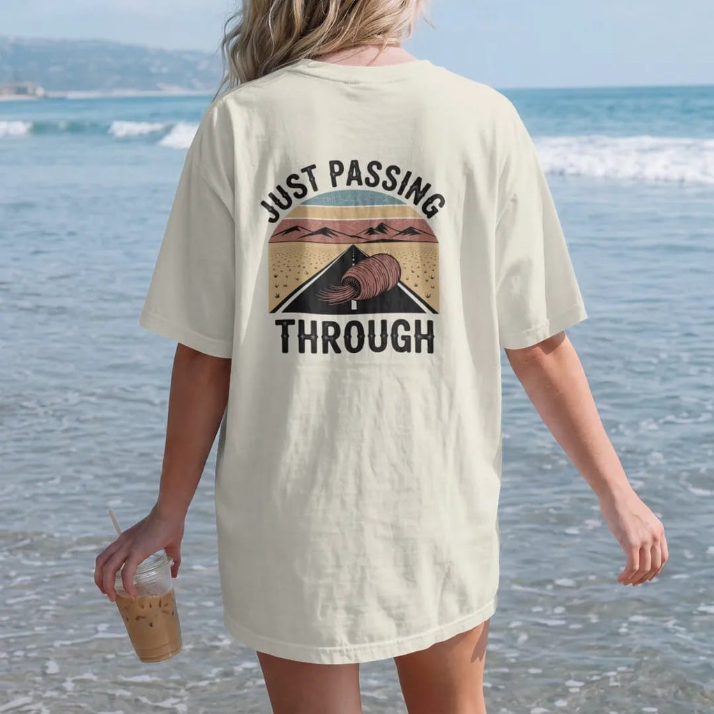 Woman on the beach in a Ivory tee, back view: a tumbleweed rolling across a flat desert road toward far mountains, "JUST PASSING THROUGH" in small retro 70s caps beneath, in a faded 1970s souvenir style with a worn cracked-ink texture, printed in…