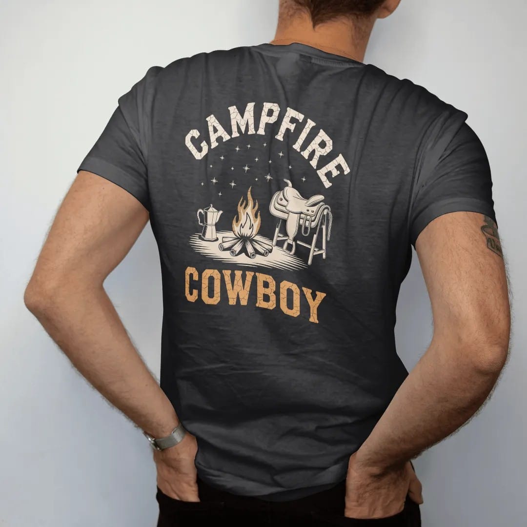 Man wearing a Black tee, seen from the back: a small campfire with a coffee pot on the coals under a sky of stars, saddle resting nearby, "CAMPFIRE" arched above and "COWBOY" below in chunky 70s slab letters, in a faded 1970s souvenir style with a…