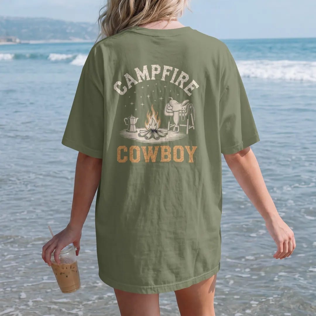 Woman on the beach in a Moss tee, back view: a small campfire with a coffee pot on the coals under a sky of stars, saddle resting nearby, "CAMPFIRE" arched above and "COWBOY" below in chunky 70s slab letters, in a faded 1970s souvenir style with a…