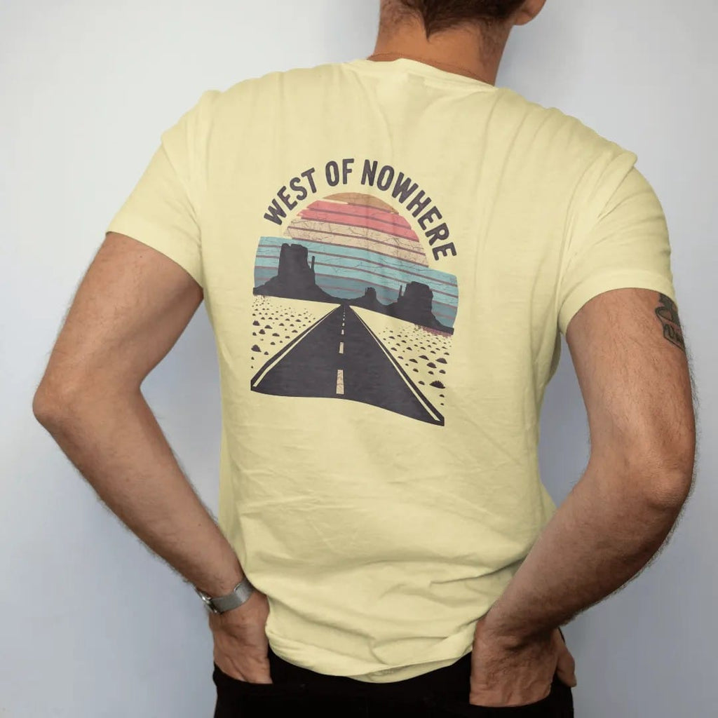 Man wearing a Butter tee, seen from the back: a straight desert highway running to a vanishing point between mesas under a retro-striped sun, "WEST OF NOWHERE" in faded 70s souvenir-sign lettering across the top, in a faded 1970s souvenir style…