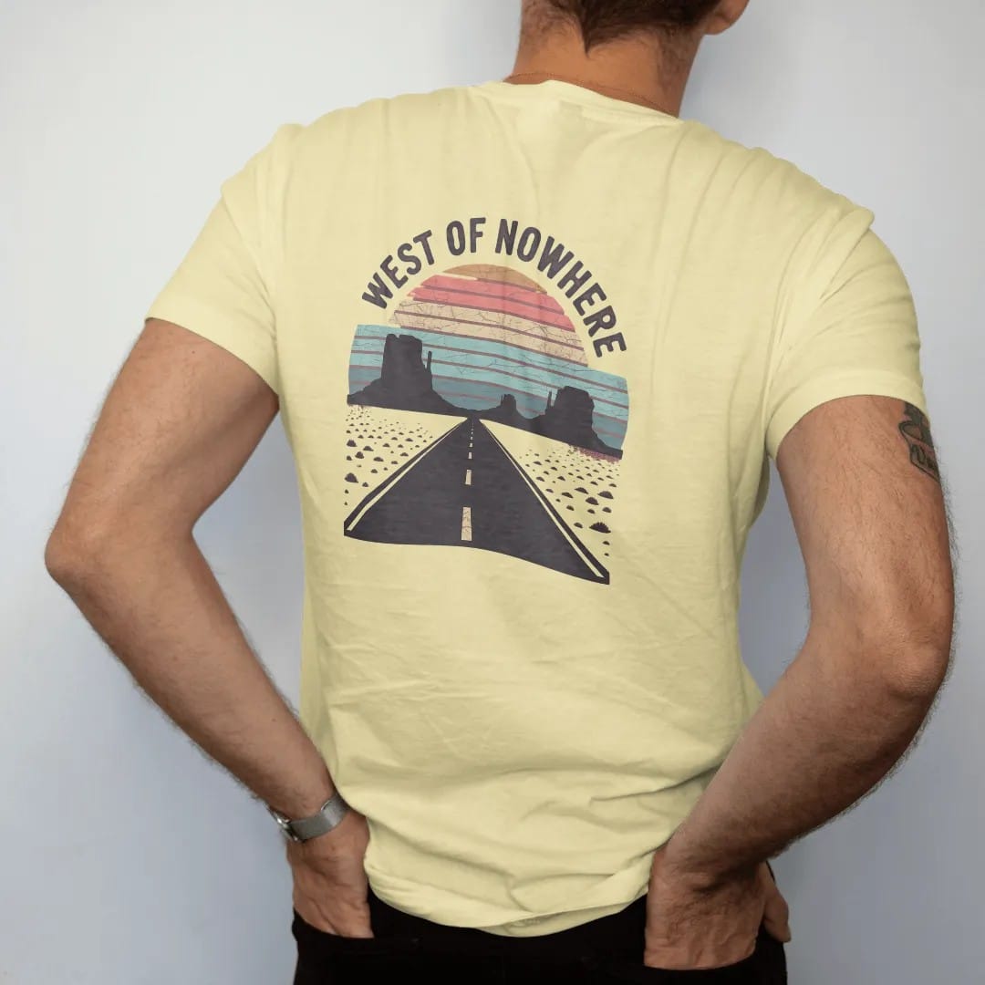 Man wearing a Butter tee, seen from the back: a straight desert highway running to a vanishing point between mesas under a retro-striped sun, "WEST OF NOWHERE" in faded 70s souvenir-sign lettering across the top, in a faded 1970s souvenir style…