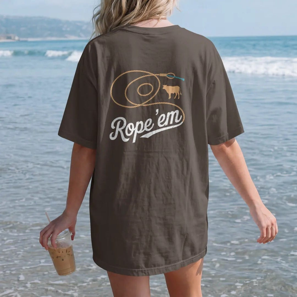 Woman on the beach in a Espresso tee, back view: a coiled lasso flying open into a big loop with a small calf silhouette, "ROPE 'EM" in a looping 70s rope script across the loop, in a faded 1970s souvenir style with a worn cracked-ink texture,…