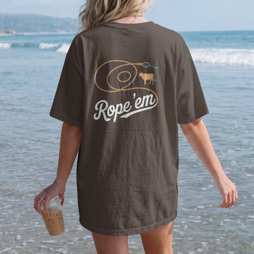 Woman on the beach in a Espresso tee, back view: a coiled lasso flying open into a big loop with a small calf silhouette, "ROPE 'EM" in a looping 70s rope script across the loop, in a faded 1970s souvenir style with a worn cracked-ink texture,…