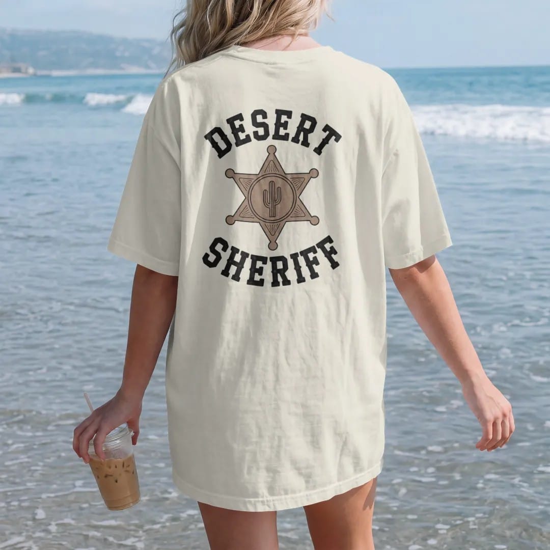 Woman on the beach in a Ivory tee, back view: a worn six-point sheriff star badge with a small cactus engraved in the center, "DESERT" in chunky 70s slab letters above and "SHERIFF" in chunky 70s block letters below, in a faded 1970s souvenir style…